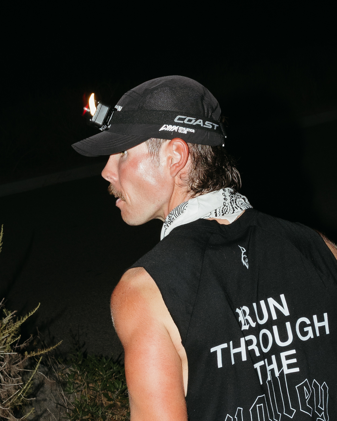 A man in profile wearing a black sleeveless shirt with 'RUN THROUGH THE VALLEY' printed in white on the back, a black lynx brand cap with a mounted headlamp on, and a white patterned neck gaiter, photographed at night outdoors.