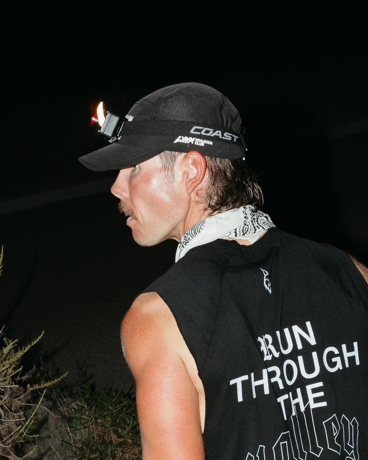 A man in profile wearing a black sleeveless shirt with 'RUN THROUGH THE VALLEY' printed in white on the back, a black lynx brand cap with a mounted headlamp on, and a white patterned neck gaiter, photographed at night outdoors.