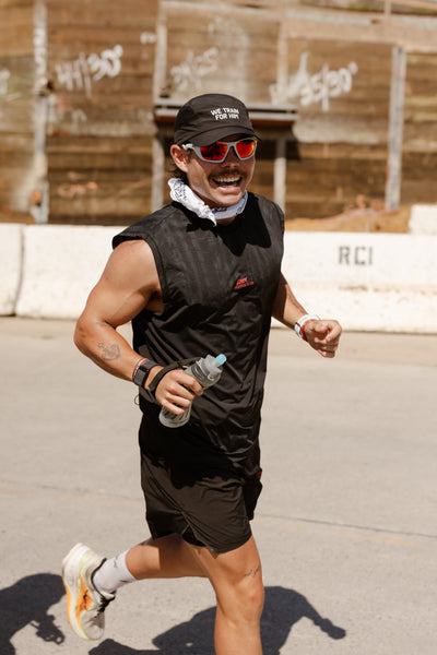 A male runner in a black sleeveless top, black shorts, and a black cap with red-tinted sunglasses is running outdoors on a paved road, holding a water bottle. A concrete retaining wall with graffiti is in the background.